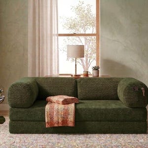 May include: A low-profile olive green corduroy sofa featuring a rectangular silhouette with rounded bolster-style armrests. A folded, multi-coloured textured throw blanket rests on the seat cushion, and a small metal key hangs from the right arm.