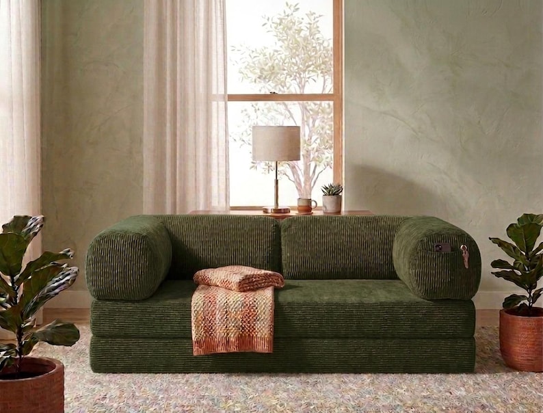May include: A low-profile olive green corduroy sofa featuring a rectangular silhouette with rounded bolster-style armrests. A folded, multi-coloured textured throw blanket rests on the seat cushion, and a small metal key hangs from the right arm.