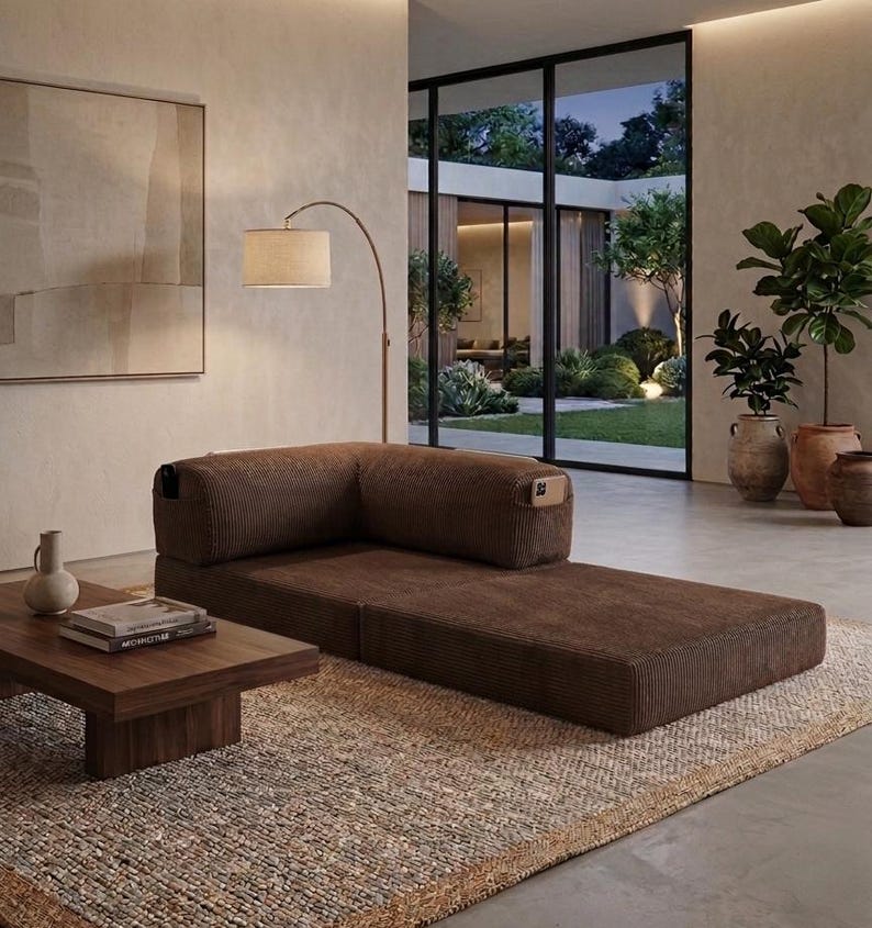 May include: A low-profile, brown corduroy modular sofa with a corner backrest and integrated side storage pockets. The sofa is positioned on a textured rug next to a low wooden coffee table, featuring a clean, rectangular design and firm cushions.