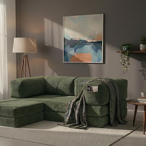 May include: A modular olive green corduroy sofa with a low-profile design and a matching ottoman. The sofa features a built-in side pocket containing a small book, and a textured grey throw blanket is draped over the right armrest.