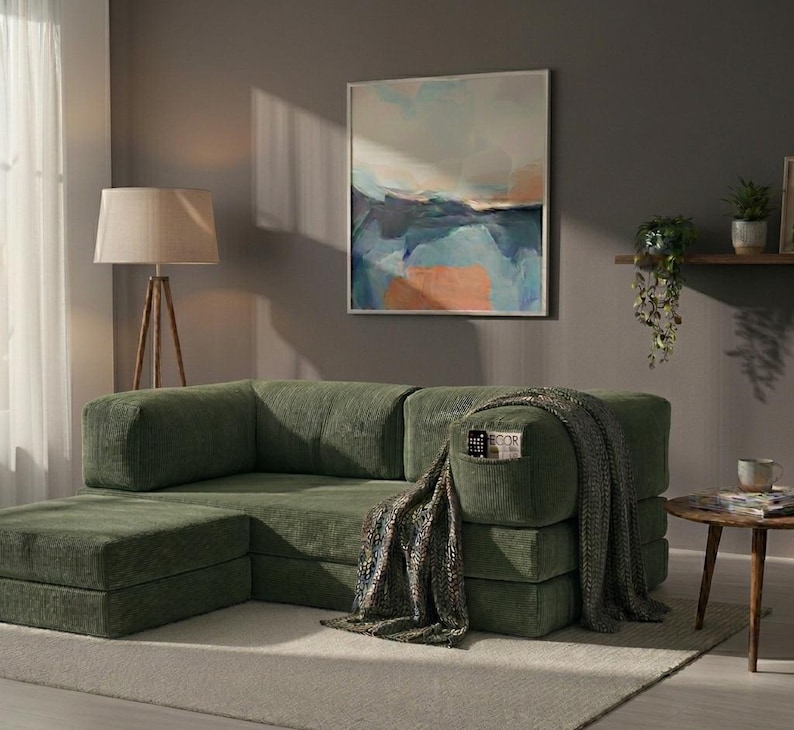 May include: A modular olive green corduroy sofa with a low-profile design and a matching ottoman. The sofa features a built-in side pocket containing a small book, and a textured grey throw blanket is draped over the right armrest.