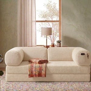 May include: A cream-colored corduroy sofa with rounded armrests and a low-profile design. A multi-colored woven throw blanket is draped over the center seat cushion. The right armrest features a small integrated side pocket containing a phone.