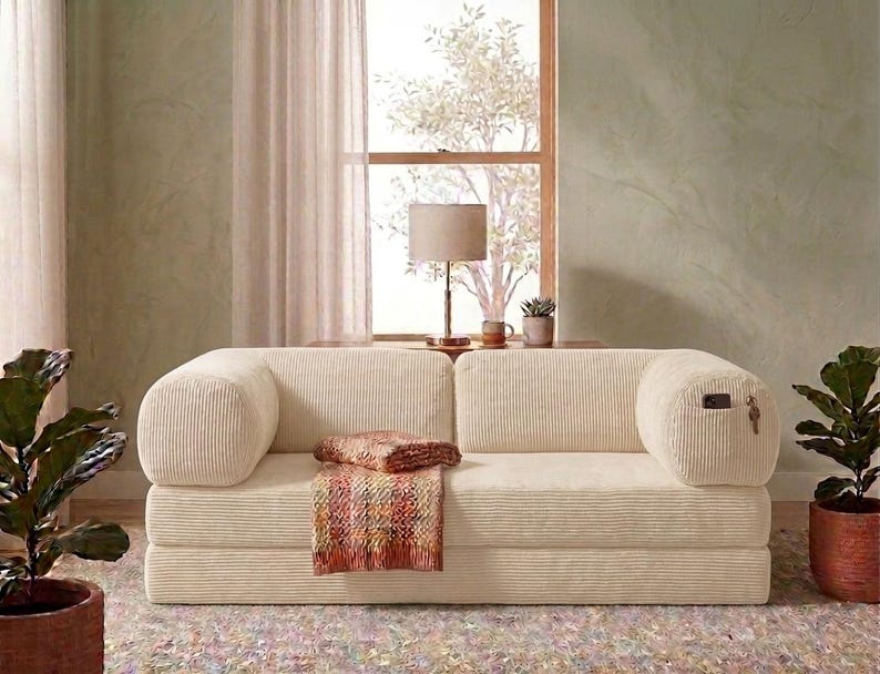 May include: A cream-colored corduroy sofa with rounded armrests and a low-profile design. A multi-colored woven throw blanket is draped over the center seat cushion. The right armrest features a small integrated side pocket containing a phone.