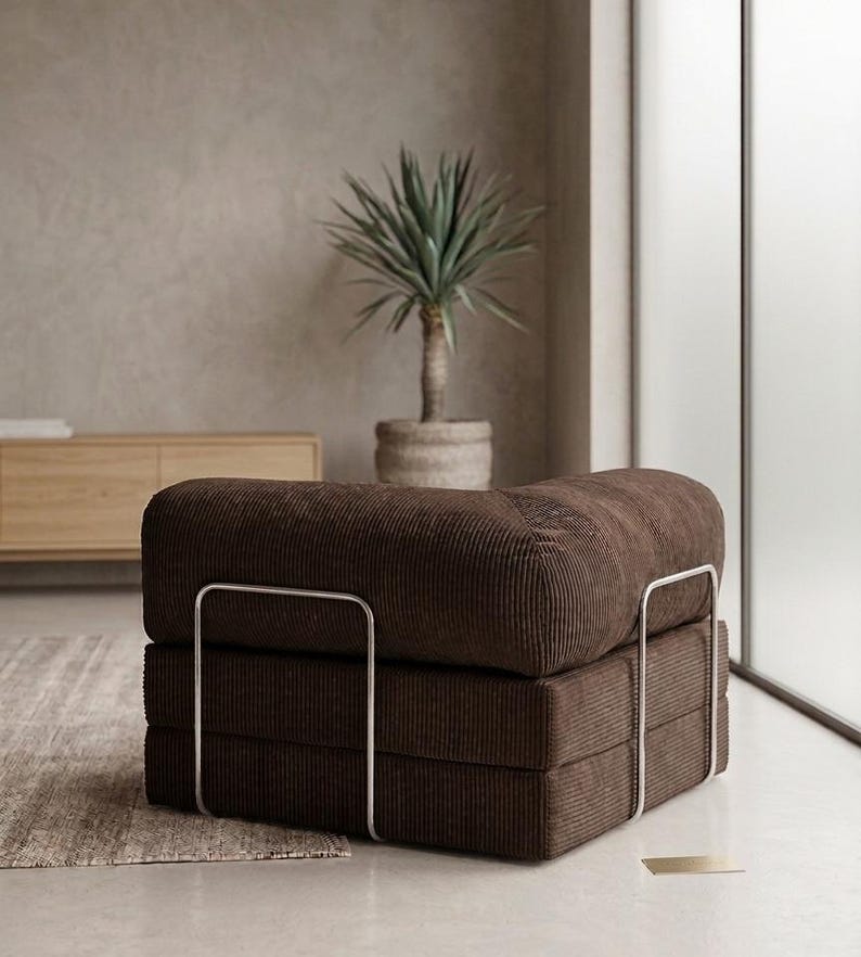 May include: A low-profile, square-shaped armchair upholstered in dark brown corduroy fabric. The seat features a thick, cushioned design supported by a sleek, silver-toned metal frame that wraps around the exterior sides of the base and backrest.