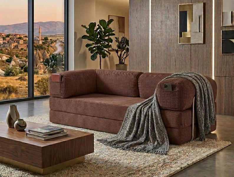 May include: A brown corduroy sofa with a low-profile design, featuring integrated side pockets on the armrests. A grey and white textured throw blanket with fringe edges is draped over the right arm, and the sofa has a structured, rectangular frame.