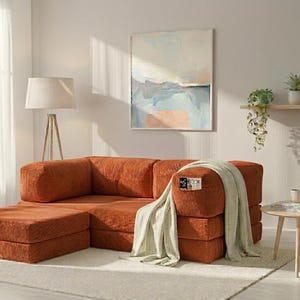 May include: A modular rust-colored corduroy sofa with a matching ottoman, draped with a light beige knit throw blanket. The sofa features a small rectangular tag with the text "ECOR" on the front armrest, set in a bright room with a floor lamp.
