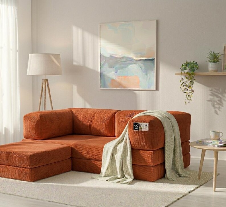 May include: A modular rust-colored corduroy sofa with a matching ottoman, draped with a light beige knit throw blanket. The sofa features a small rectangular tag with the text "ECOR" on the front armrest, set in a bright room with a floor lamp.