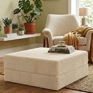 May include: A square, cream-colored corduroy ottoman sits in front of an upholstered armchair. The chair features a textured cream fabric and is draped with a mustard yellow knit throw blanket. A stack of books sits atop the ottoman.