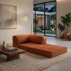 May include: A low-profile, rust-colored corduroy modular sofa with an L-shaped backrest and integrated side pockets. The sofa is positioned on a textured woven rug next to a dark wood coffee table holding a ceramic vase and stacked books.