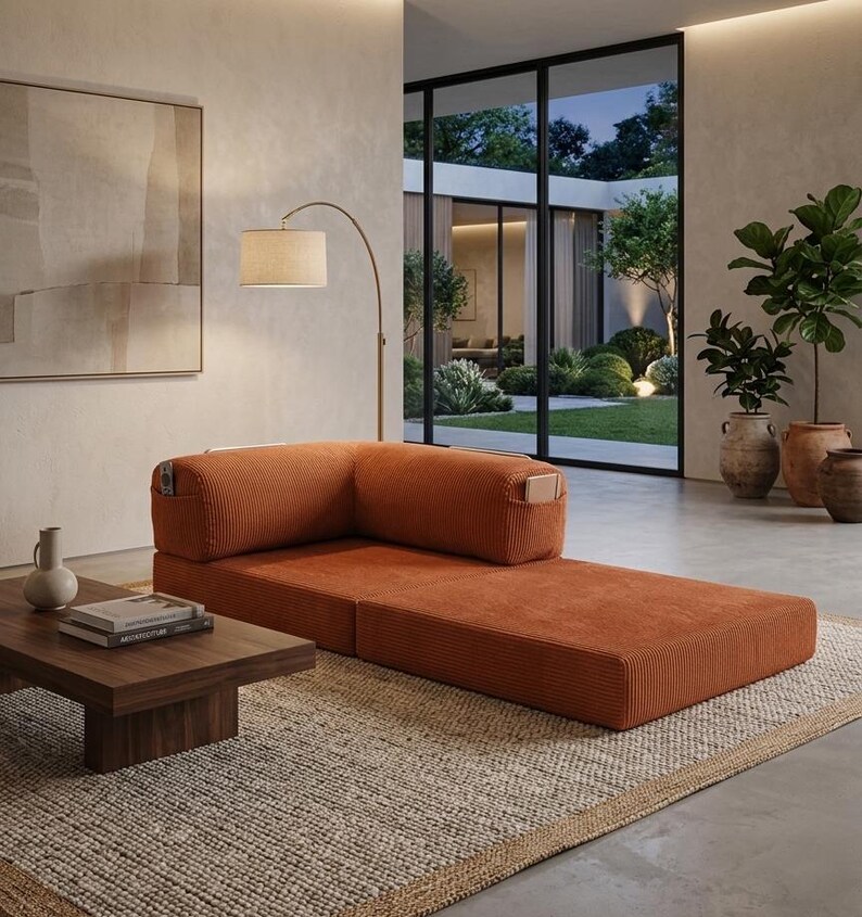 May include: A low-profile, rust-colored corduroy modular sofa with an L-shaped backrest and integrated side pockets. The sofa is positioned on a textured woven rug next to a dark wood coffee table holding a ceramic vase and stacked books.