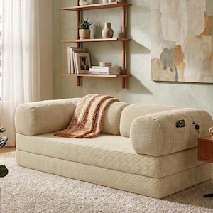 May include: A cream-colored corduroy sofa bed featuring a low-profile design with rounded armrests. The sofa includes a side pocket containing a smartphone and keys, and a textured orange and white throw blanket draped over the center seat cushion.