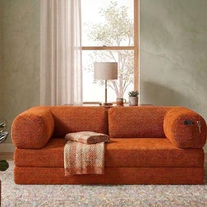 May include: A burnt orange corduroy sofa featuring a low-profile design with rounded armrests, a thick seat cushion, and a matching textured throw blanket draped over the center. The right armrest includes a small side pocket containing a key.