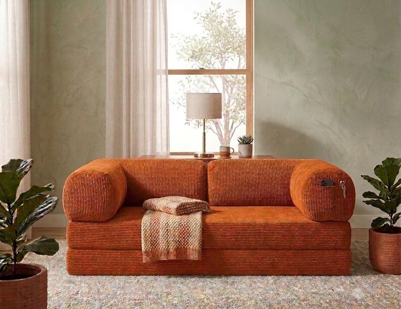 May include: A burnt orange corduroy sofa featuring a low-profile design with rounded armrests, a thick seat cushion, and a matching textured throw blanket draped over the center. The right armrest includes a small side pocket containing a key.