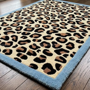 May include: A rectangular area rug featuring a cream-colored base with a repeating black and brown leopard print pattern. The rug is finished with a solid light blue border around the entire perimeter. The text ETHICAL THREADS is visible.