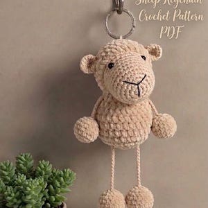 May include: A beige crochet sheep keychain hanging from a metal ring. The sheep has a round head, small ears, a round body, and two long legs ending in round feet. The text "Sheep Keychain Crochet Pattern PDF" is visible in the upper right corner.