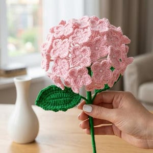 May include: A handmade crochet hydrangea flower in shades of pink, green, and white. The flower has a detailed, textured appearance with a green stem and leaf. The flower is held in a person's hand.