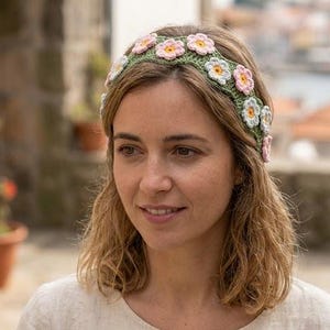 May include: A crochet headband featuring a floral design. The headband is green with pink, white, and yellow flowers. The flowers are evenly spaced around the band. The woman in the image is wearing the headband.