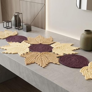 May include: A crochet table runner composed of interconnected light yellow maple leaf shapes and dark purple circular motifs. The textile piece features a repeating pattern of leaves and circles, creating a decorative arrangement for a flat surface.