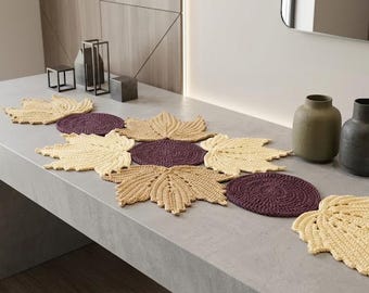 May include: A crochet table runner composed of interconnected light yellow maple leaf shapes and dark purple circular motifs. The textile piece features a repeating pattern of leaves and circles, creating a decorative arrangement for a flat surface.