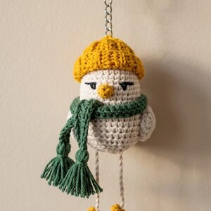 May include: A crocheted bird keychain featuring a cream-colored body, a mustard yellow beanie, a dark green scarf with tassels, and yellow feet. The bird has black embroidered eyes and a small yellow beak, attached to a silver metal keyring and chain.