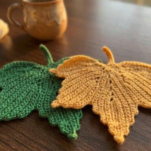 Può includere: Due sottobicchieri all'uncinetto a forma di foglia, uno di un verde vivace e l'altro di un caldo giallo dorato. Ogni sottobicchiere presenta una superficie dettagliata e strutturata con uno stelo centrale e venature definite come una foglia.