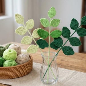 May include: Three crochet leaf stems in varying shades of green, ranging from light lime to dark forest green, are arranged in a clear glass vase. Beside the vase sits a round woven basket filled with several balls of green and cream yarn.