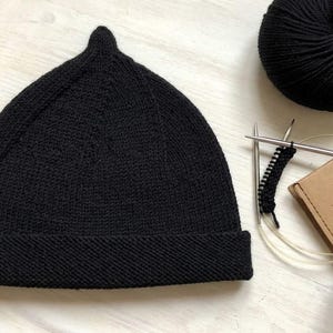 Puede incluir: Un gorro de punto negro liso con una corona puntiaguda y un ala ancha doblada. Junto al gorro hay dos ovillos de lana negra, un par de agujas de tejer circulares y una pequeña parte de lana negra trabajándose en las agujas.