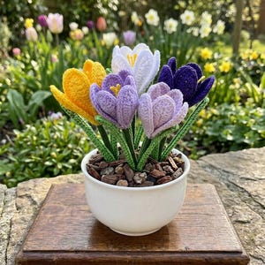 May include: A handcrafted crochet flower arrangement featuring yellow, purple, white, and dark purple crocus flowers. The flowers are in a white ceramic pot filled with wood chips, sitting on a wooden base. The background shows a garden with other flowers.