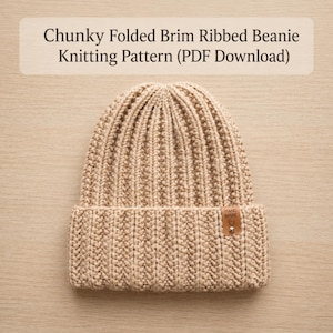 Puede incluir: Un gorro beige de punto grueso acanalado con borde doblado y una pequeña etiqueta rectangular marrón con la inscripción "HAND MADE" y un remache plateado. El texto "Chunky Folded Brim Ribbed Beanie Knitting Pattern (PDF Download)" aparece arriba.