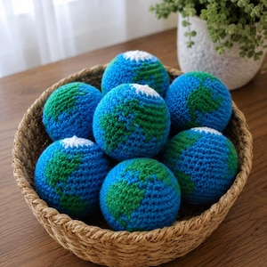 May include: A woven basket filled with seven crocheted balls, each depicting the Earth. The balls are primarily blue with green and white accents, representing continents and polar regions. The basket sits on a wooden surface.