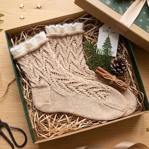 May include: A pair of beige knit socks with a delicate lace pattern and a soft white cuff, nestled inside a gift box filled with shredded paper, a pinecone, cinnamon sticks, and a gift tag labeled with a tree and the text With Love.
