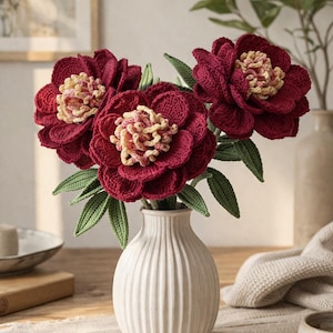 Op de afbeelding: Een boeket van drie handgemaakte gehaakte bloemen in een witte vaas. De bloemen zijn diep bordeauxrood met gele en roze centra en groene bladeren. De vaas heeft verticale ribbels.
