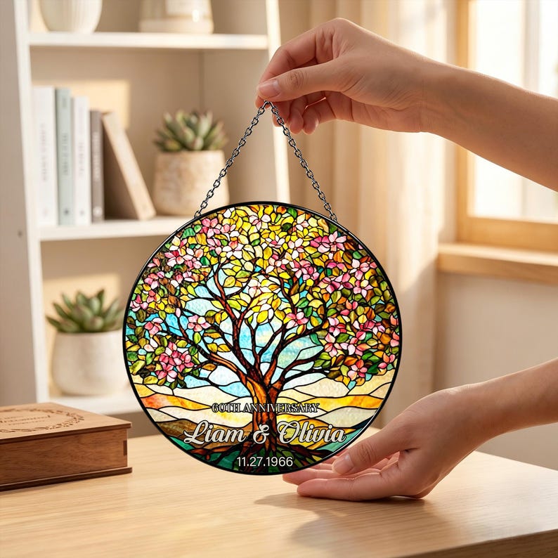 May include: A round stained glass window with a tree design. The tree has pink and yellow flowers, with a blue sky background. The text "60th Anniversary" and the names "Liam & Olivia" are displayed, along with the date 11.27.1966.