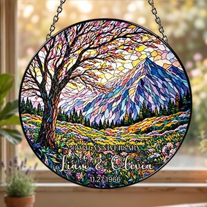 May include: A round stained glass window with a colorful landscape design. The image features a tree, mountains, and a field of flowers. Text reads "60th Anniversary" and "Liam & Olivia" with the date "11.27.1966". The window is framed in black and hangs from a chain.