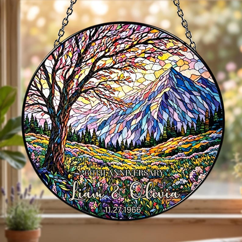 May include: A round stained glass window with a colorful landscape design. The image features a tree, mountains, and a field of flowers. Text reads "60th Anniversary" and "Liam & Olivia" with the date "11.27.1966". The window is framed in black and hangs from a chain.