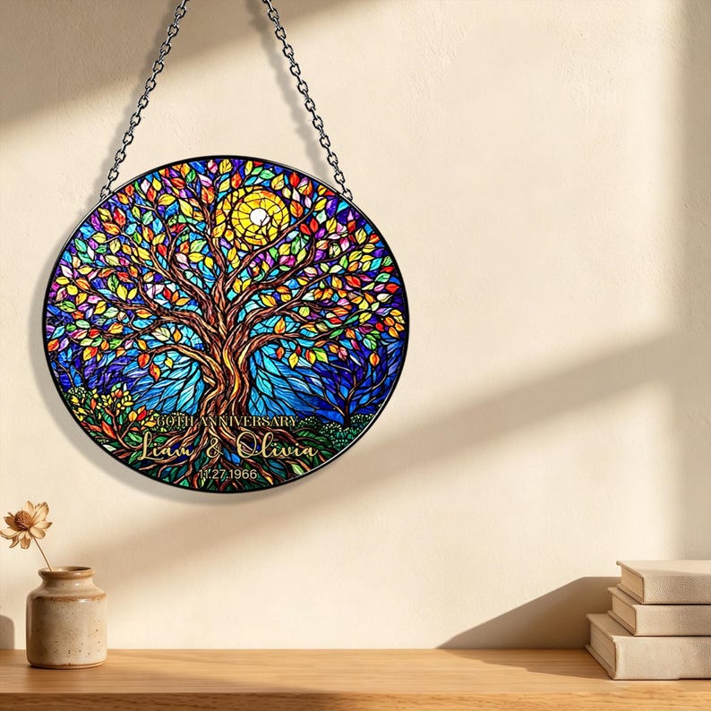 May include: A round stained glass panel featuring a colorful tree with a yellow sun. The tree has vibrant leaves in shades of red, orange, yellow, and green. The text "Liam & Olivia" and "11.27.1966" is displayed on the bottom. The panel is hanging from a chain.