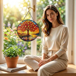 May include: A round stained glass window hanging from a gold hook. The window features a tree with red and orange leaves, a lake, and the text "Liam & Olivia 1.27.1966". A woman sits in front of a window with a plant in a terracotta pot.