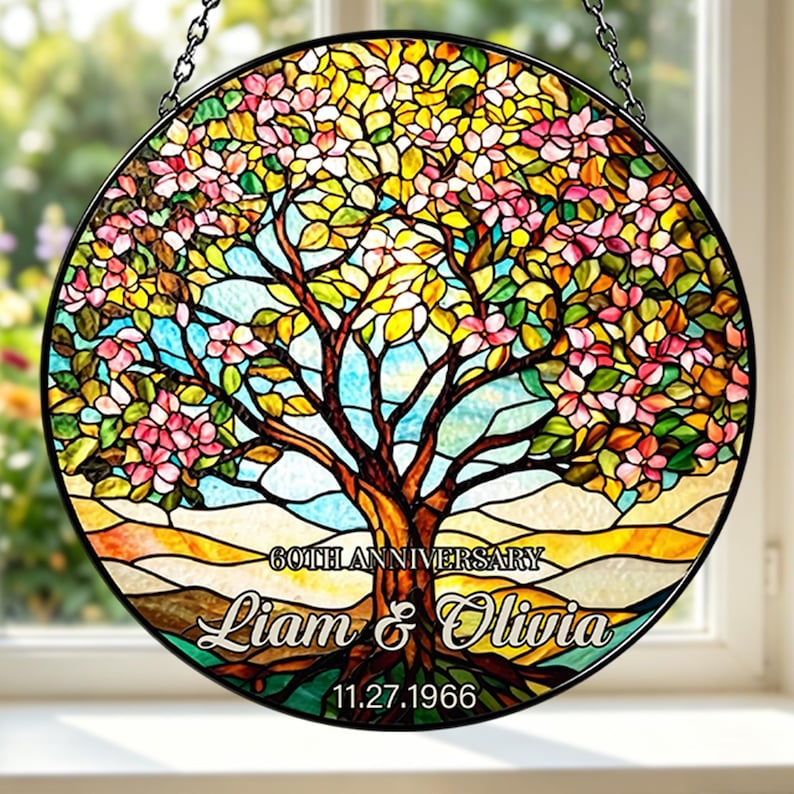 May include: A round stained glass panel featuring a tree with pink and yellow flowers, brown trunk, and green leaves. The text "60th Anniversary" is above the names "Liam & Olivia" and the date "11.27.1966". The panel is framed in black and hangs from a chain.