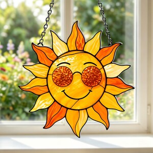 Op de afbeelding: Een glas-in-lood zonnevanger met een felgeel en oranje gezicht met een glimlachende uitdrukking en een ronde zonnebril van getextureerd barnsteenkleurig glas, opgehangen aan een zilverkleurige metalen ketting voor een raam.