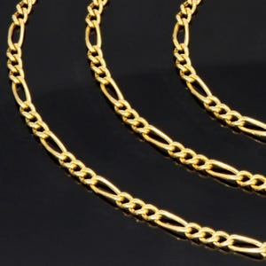 K&ouml;nnte beinhalten: Eine goldfarbene Figaro-Kette mit einem sich wiederholenden Muster aus drei kleinen runden Gliedern, gefolgt von einem l&auml;nglichen ovalen Glied. Die metallische Kette hat eine polierte Oberfl&auml;che und ist in mehreren geschwungenen Schleifen angeordnet.