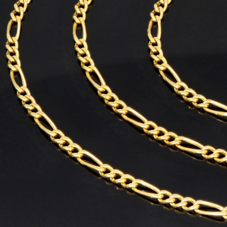 K&ouml;nnte beinhalten: Eine goldfarbene Figaro-Kette mit einem sich wiederholenden Muster aus drei kleinen runden Gliedern, gefolgt von einem l&auml;nglichen ovalen Glied. Die metallische Kette hat eine polierte Oberfl&auml;che und ist in mehreren geschwungenen Schleifen angeordnet.