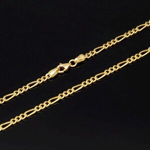 K&ouml;nnte beinhalten: Eine goldfarbene Figaro-Halskette mit einem sich wiederholenden Muster aus drei kleinen runden Gliedern, gefolgt von einem l&auml;nglichen ovalen Glied. Die Kette wird mit einem polierten Karabinerverschluss in der Mitte des Bildes geschlossen.