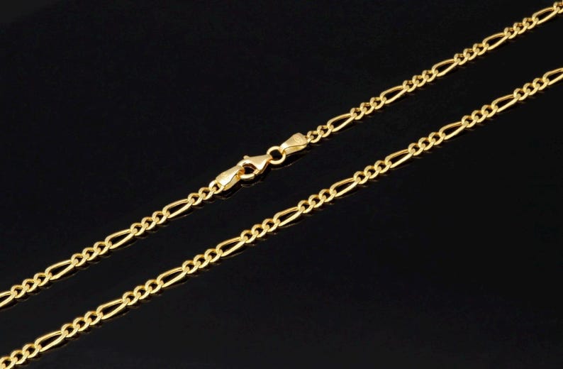 K&ouml;nnte beinhalten: Eine goldfarbene Figaro-Halskette mit einem sich wiederholenden Muster aus drei kleinen runden Gliedern, gefolgt von einem l&auml;nglichen ovalen Glied. Die Kette wird mit einem polierten Karabinerverschluss in der Mitte des Bildes geschlossen.