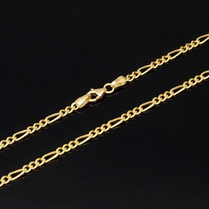 K&ouml;nnte beinhalten: Eine goldfarbene Figaro-Halskette mit einem sich wiederholenden Muster aus drei kleinen runden Gliedern, gefolgt von einem l&auml;nglichen ovalen Glied. Die Kette wird mit einem Karabinerverschluss geschlossen, der in der Bildmitte zu sehen ist.