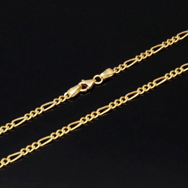 K&ouml;nnte beinhalten: Eine goldfarbene Figaro-Halskette mit einem sich wiederholenden Muster aus drei kleinen runden Gliedern, gefolgt von einem l&auml;nglichen ovalen Glied. Die Kette wird mit einem Karabinerverschluss geschlossen, der in der Bildmitte zu sehen ist.