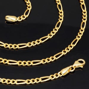 K&ouml;nnte beinhalten: Eine goldfarbene Figaro-Halskette mit einem sich wiederholenden Muster aus drei kleinen runden Gliedern, gefolgt von einem l&auml;nglichen ovalen Glied. Die Kette ist an einem Ende mit einem Karabinerverschluss und am anderen mit einer Metallplatte versehen.