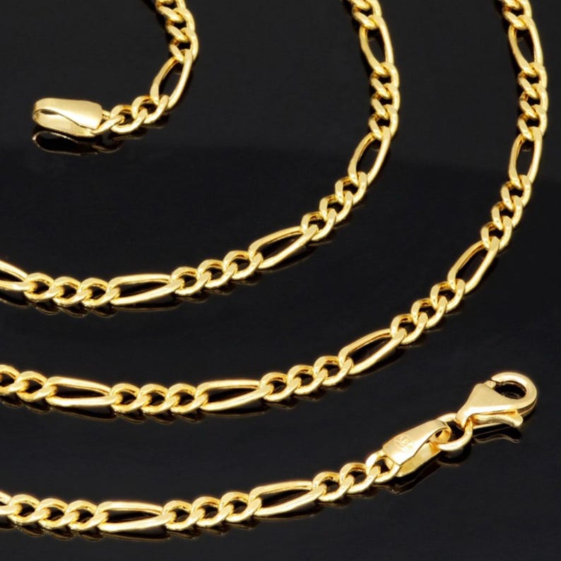 K&ouml;nnte beinhalten: Eine goldfarbene Figaro-Halskette mit einem sich wiederholenden Muster aus drei kleinen runden Gliedern, gefolgt von einem l&auml;nglichen ovalen Glied. Die Kette ist an einem Ende mit einem Karabinerverschluss und am anderen mit einer Metallplatte versehen.