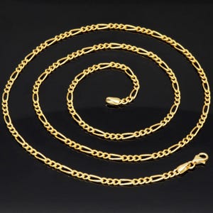 K&ouml;nnte beinhalten: Eine goldfarbene Figaro-Halskette mit einem sich wiederholenden Muster aus drei kleinen runden Gliedern, gefolgt von einem l&auml;nglichen ovalen Glied. Die Kette ist spiralf&ouml;rmig angeordnet und verf&uuml;gt &uuml;ber einen sicheren Karabinerverschluss.