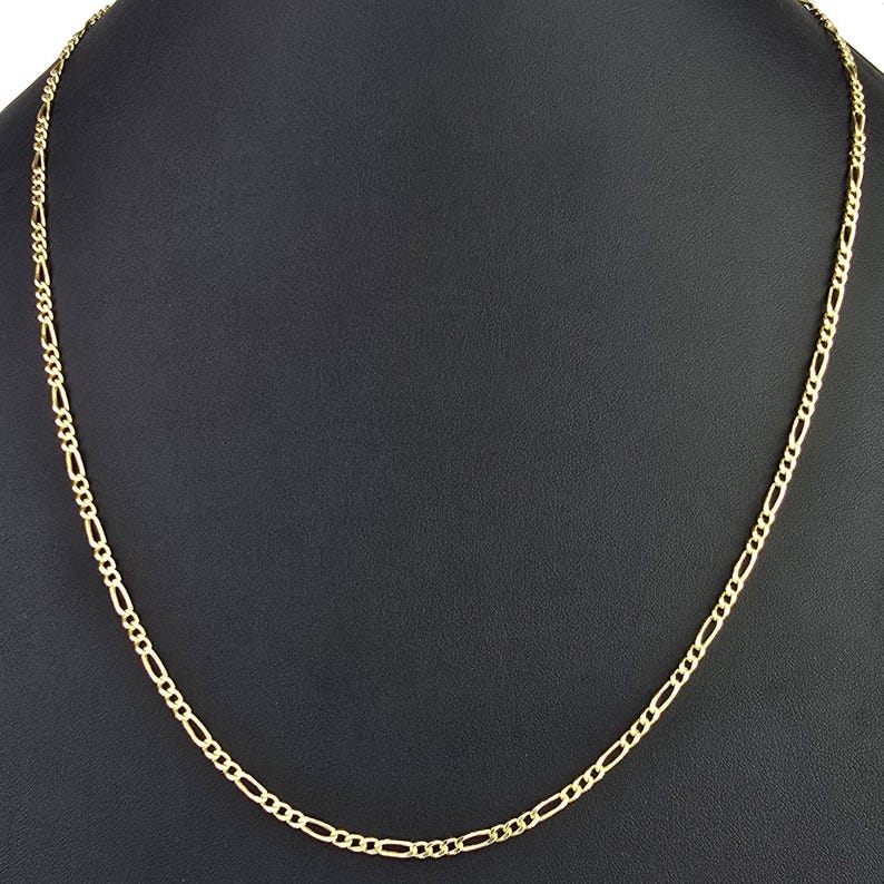 K&ouml;nnte beinhalten: Eine goldfarbene Metallkettenhalskette mit einem klassischen Figaro-Gliedermuster. Die Kette besteht aus einem sich wiederholenden Muster aus drei kleinen runden Gliedern, gefolgt von einem l&auml;nglichen ovalen Glied, das einen gleichm&auml;&szlig;igen Strang bildet.