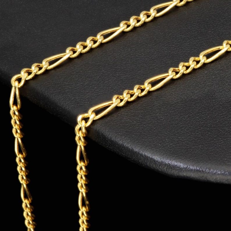 K&ouml;nnte beinhalten: Eine goldfarbene Figaro-Kette mit einem sich wiederholenden Muster aus drei kleinen runden Gliedern, gefolgt von einem l&auml;nglichen ovalen Glied. Die Metallkette hat eine polierte Oberfl&auml;che und ist in einer winkligen Form drapiert.